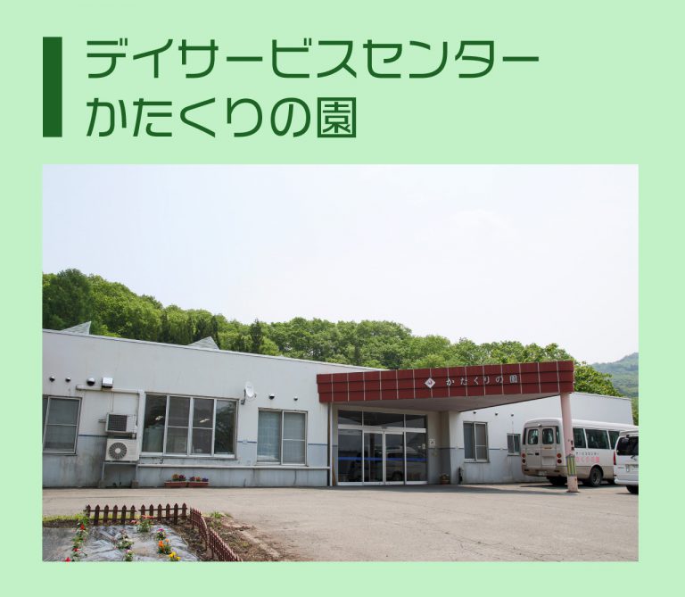 デイサービスセンター かにくりの園の建物外観。山を背景に白い施設と赤い看板が見える。